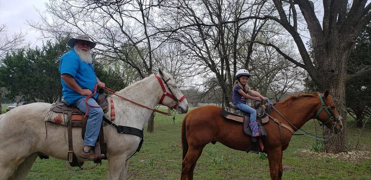 A man in a cowboy hat on a gray horse and a young girl in a riding helmet on a chestnut horse, ready for a trail ride in the Texas Hill Country