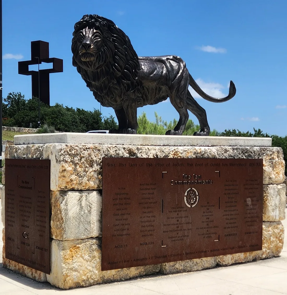 Bronze Lion of Judah statue on a limestone base with the tall rust-colored steel cross rising behind it at the Coming King Sculpture Prayer Garden in Kerrville, Texas
